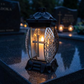 Znicz kapliczka czarno-srebrny ze srebrnym krzyżem zapalony na nagrobku nocą